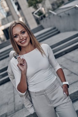 Güzel ve başarılı, kameraya bakarak ve açık havada duran süre gülümseyen elbiseli çekici genç kadın