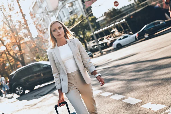 blonde young woman in suit pulling luggage while walking outdoors in city street road