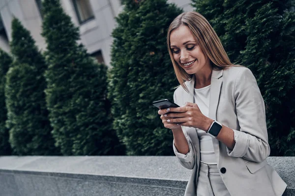 Hermosa Mujer Joven Traje Usando Teléfono Inteligente Sonriendo Mientras Está —  Fotos de Stock