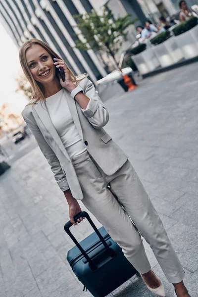 beautiful young woman in suit pulling luggage and smiling while walking outdoors and talking on mobile phone