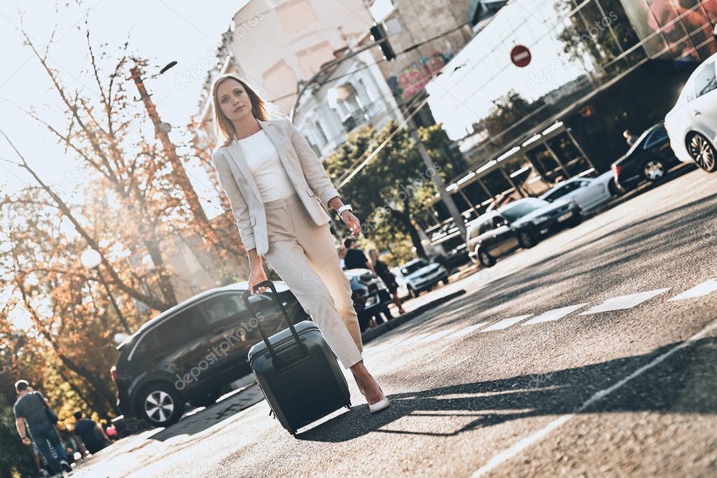 Confident businesswoman in suit pulling luggage and walking outdoors in city road street with cars 