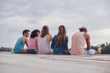 Arkadan görünüm gülümseyen ve bira şişeleri ile pier üzerine konuşurken rahat kıyafet içinde gençlerin 