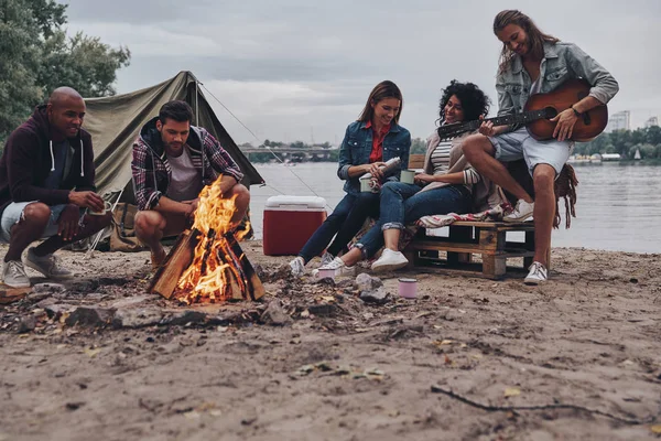 キャンプファイヤーの近くのビーチ パーティーを楽しみながら笑みを浮かべてカジュアルな服装の若い人たちのグループ — ストック写真