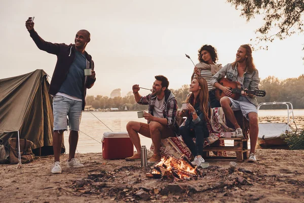 最高の携帯電話の湖と撮影 Selfie 近く休んでいる間キャンプファイヤーでマシュマロを焙煎の友人 — ストック写真