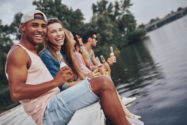 Gelukkige Jonge Mensen Vrijetijdskleding Glimlachend Houden Van Bierflesjes Zittend Pier — Stockfoto