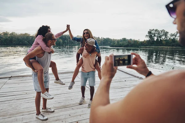 beautiful young couples spending carefree time while standing on pier, man taking mobile photo of friends having fun with piggyback