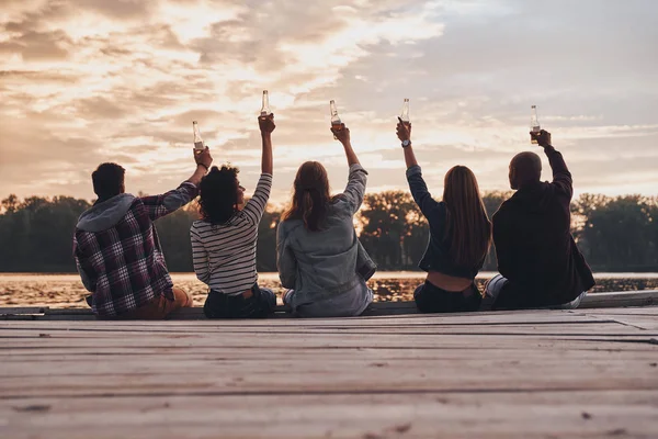 木製の桟橋の上に座って ビール瓶と乾杯の友人の背面図 — ストック写真