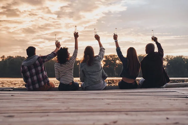 Back View Friends Sitting Wooden Pier Toasting Beer Bottles — Stock Photo, Image