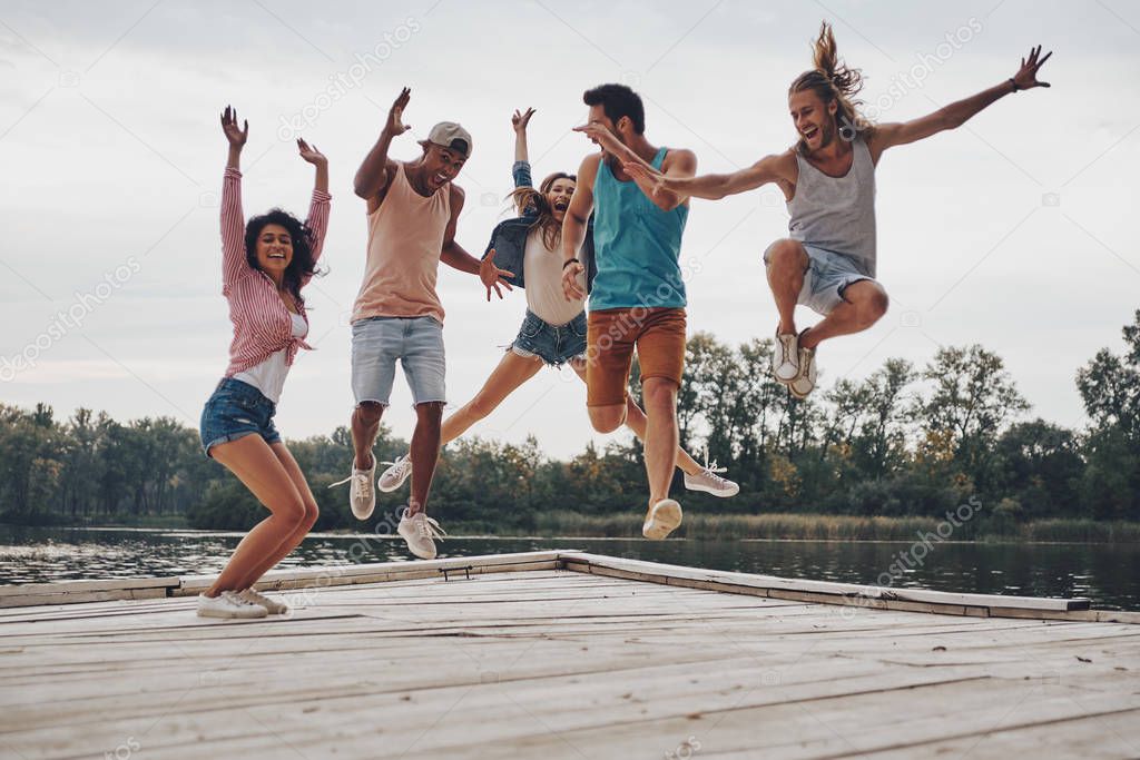 full length of young people in casual wear jumping on wooden pier at lake with forest 