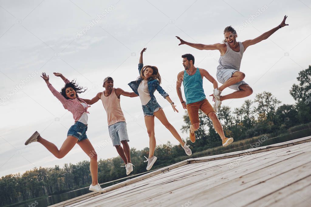 full length of young people in casual wear jumping on wooden pier at lake with forest 