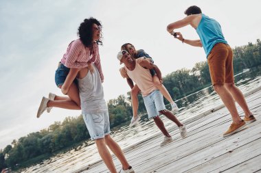 mutlu çiftler kaygısız vakit ahşap iskele üzerinde omuz kadınlar üzerinde taşıyan ve Mobil fotoğraf çekici erkek 
