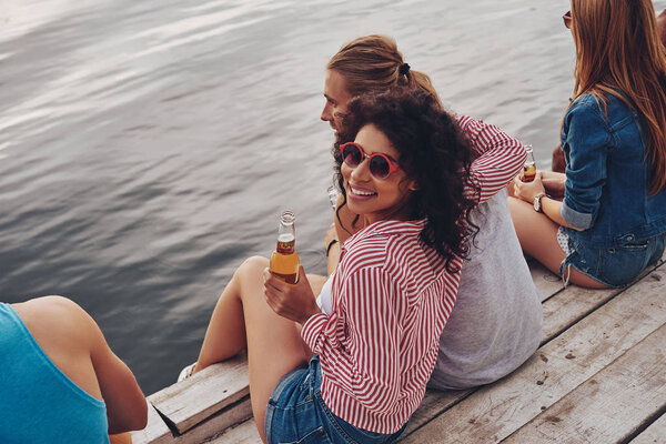 laughing young people in casual wear smiling and holding beer while sitting on pier at lake water 