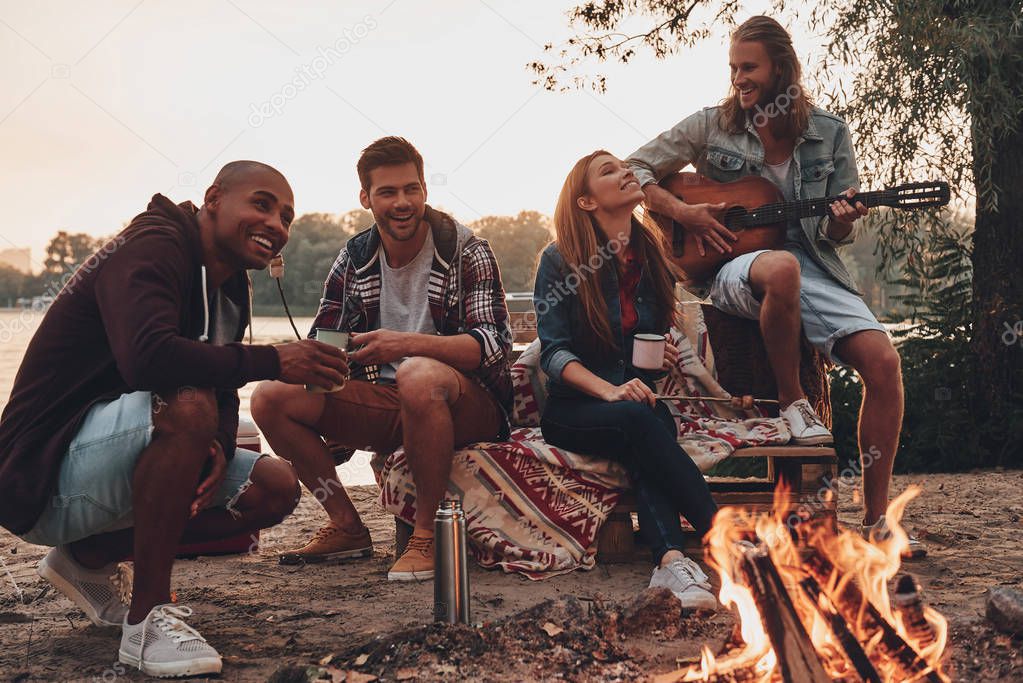 young people in casual wear enjoying camping near lake at sunset, man playing guitar 