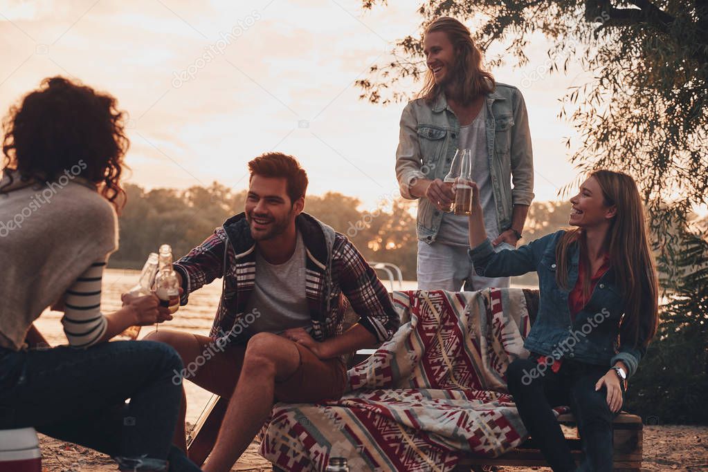group of young people in casual wear smiling and clinking with beer bottles while camping at lake