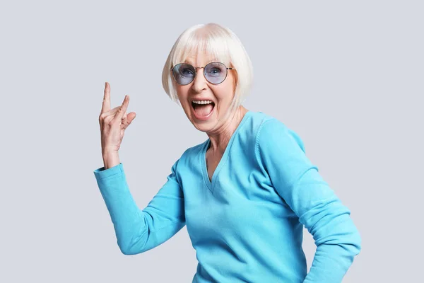 ポーズ スタジオの灰色の背景に灰色髪の短いクールな年配の女性 スタイリッシュなメガネを着用し ジェスチャー ロックの手でサイン — ストック写真