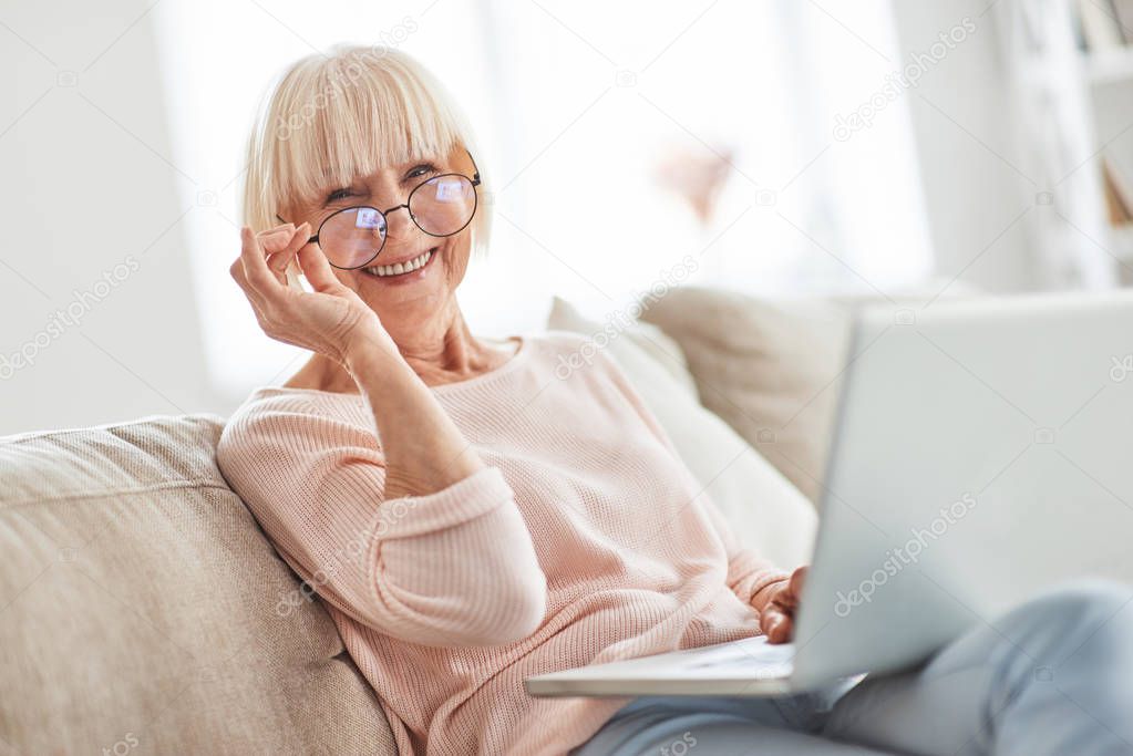 senior caucasian woman sitting on sofa at home, holding laptop on knees and looking at camera with smile 