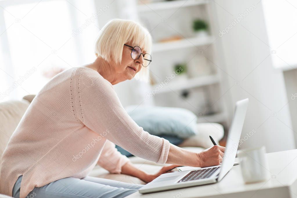 senior caucasian woman sitting on sofa at home near table with laptop and writing notes in diary