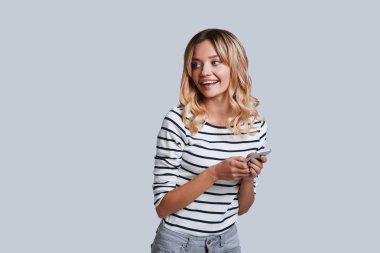 gülümseyen ve gri arka plan karşı dururken akıllı telefon tutarak sarışın genç kadın