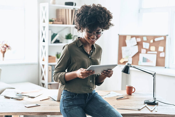 smiling young African woman using digital tablet in office room with table and lamp, book rack on background 