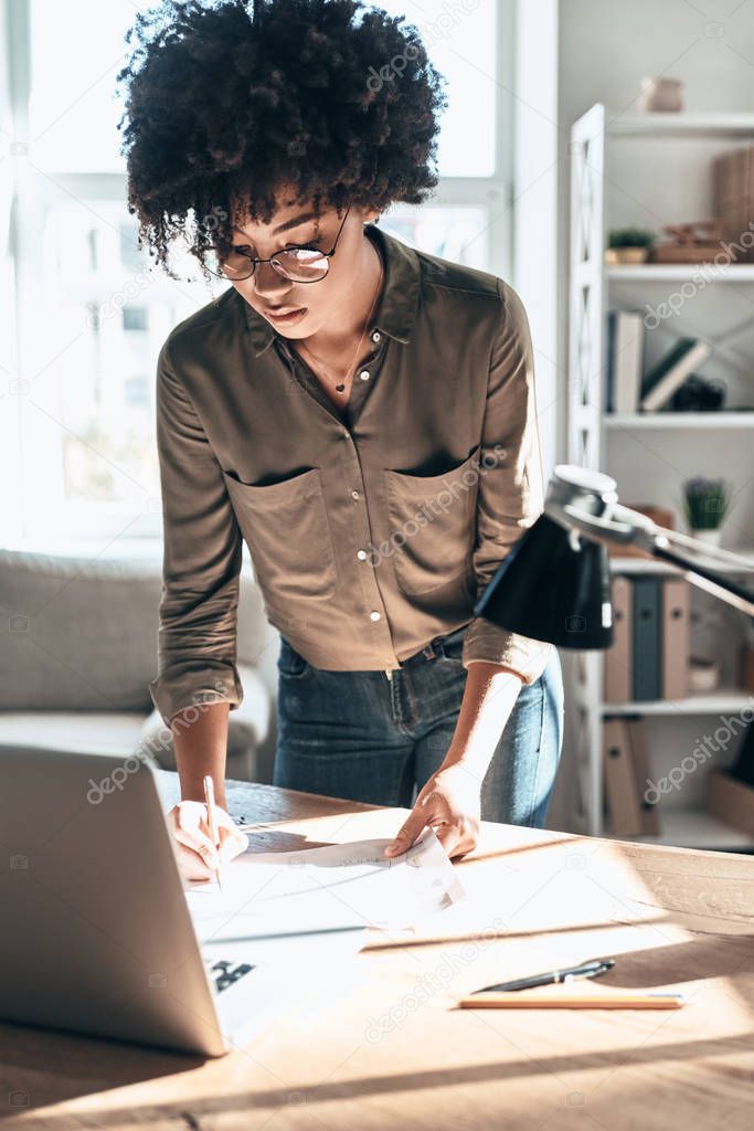 curly hair young African woman working in office at wooden table with laptop and writing 