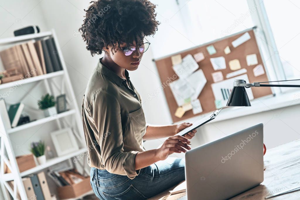serious young African woman using digital tablet and laptop ta table in office 
