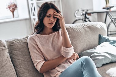 Baş ağrısı acı ve evde koltukta oturan genç kadın kötü duygu, hayal kırıklığına uğramış    