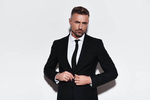Красивый молодой человек в полном костюме застегивает пиджак и смотрит в камеру, стоя на белом фоне
