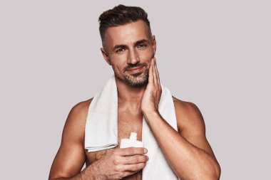 tıraş losyonu uygulamak ve omuzlarında havlu ile gri arka plan karşı dururken gülümseyen yakışıklı delikanlı 