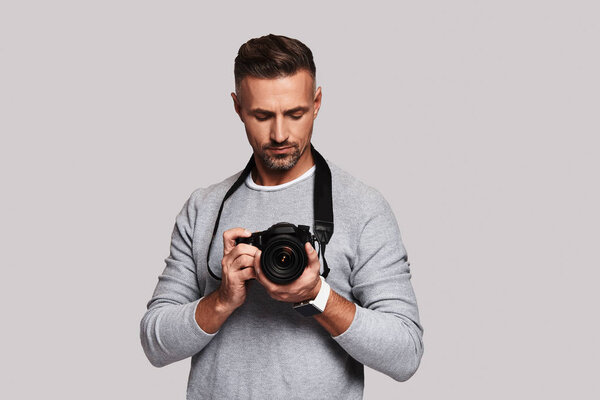 Confident photographer holding digital camera while standing against grey background