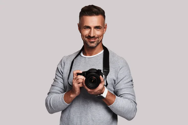 Creative good looking young man holding digital camera and smiling while standing against grey background