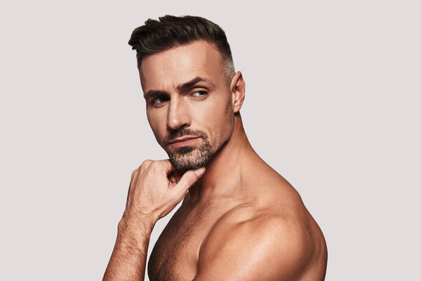 Портрет мужественности, очаровательный молодой человек держа руку на подбородке и глядя в сторону, стоя на сером фоне
