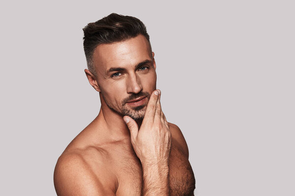 Handsome man keeping hand on chin and looking at camera while standing against grey background