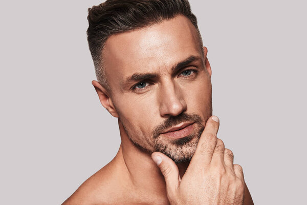 Настоящий мачо держит руку на подбородке и смотрит в камеру, стоя на сером фоне.
