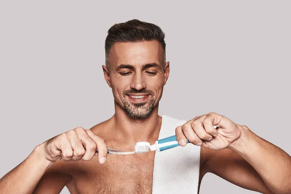 歯ブラシと灰色の背景に対して立っている間の歯磨き粉を適用するハンサムな若い男に彼の歯の世話 — ストック写真