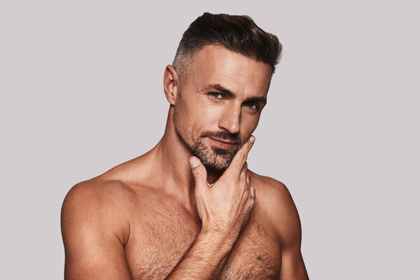 Очаровательный молодой человек держит руку на подбородке и смотрит в камеру, стоя на сером фоне
