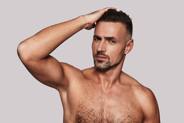 Без рубашки красивый мужчина держит руку в волосах и улыбается стоя на сером фоне
