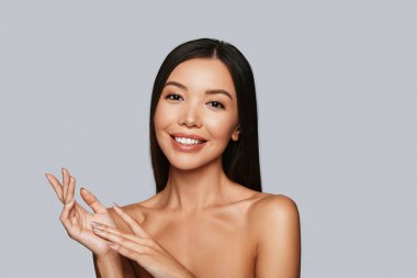 beautiful young shirtless Asian woman looking at camera and smiling while standing against grey background