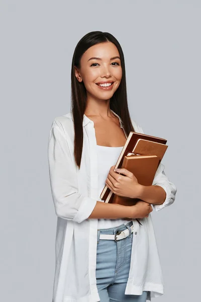 美しい若いアジア女性の本を運ぶと灰色の背景に対して立っている笑顔 — ストック写真