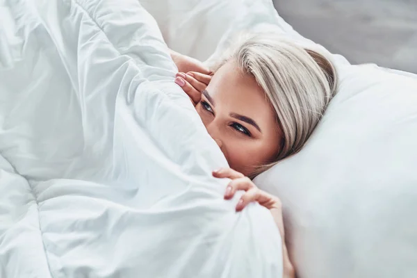Привлекательная Молодая Женщина Покрывает Половину Лица Одеялом Улыбается Лежа Дома — стоковое фото