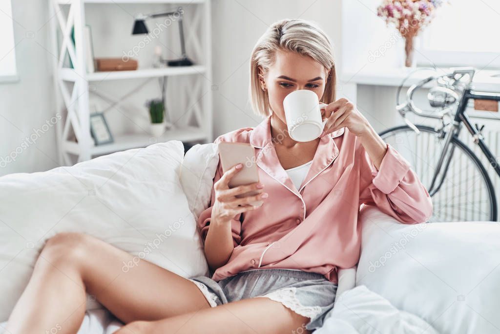 Attractive young woman using smart phone while sitting on bed at home                      