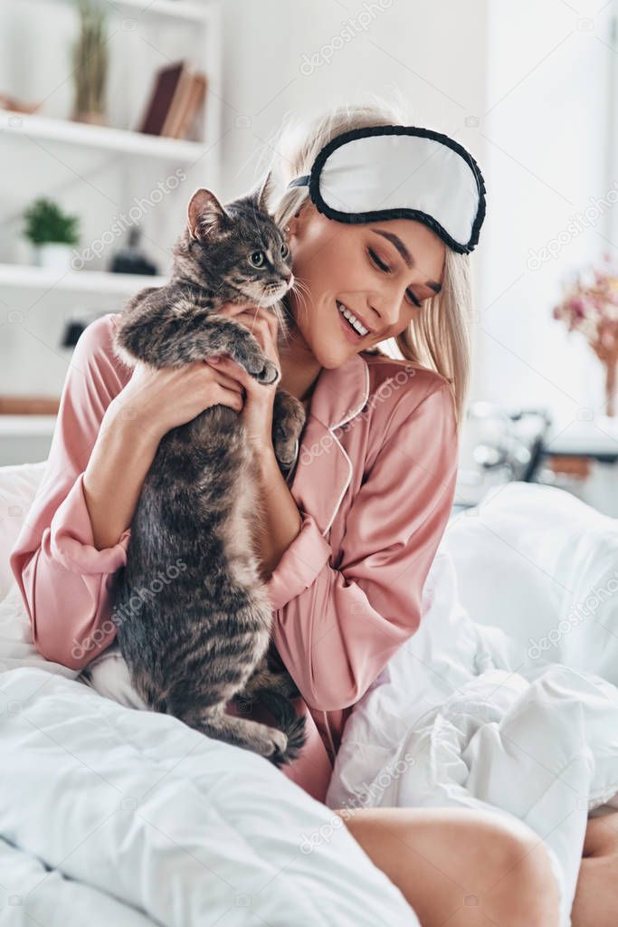 Attractive young woman playing with her cat and smiling while sitting in bed at home