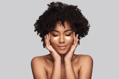 Cilt Bakımı iyi alıyor. Tıbbi göz bandı uygulamak ve gri arka plan karşı dururken gülümseyen çekici genç Afrika kadın