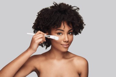 Güzel duygu. Makyaj fırça uygulamak ve gri arka plan karşı dururken gülümseyen çekici genç Afrika kadın