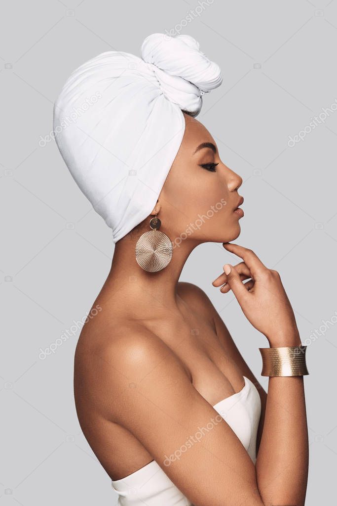 African queen. Side view of attractive young African woman in turban keeping hand on chin while standing against grey background