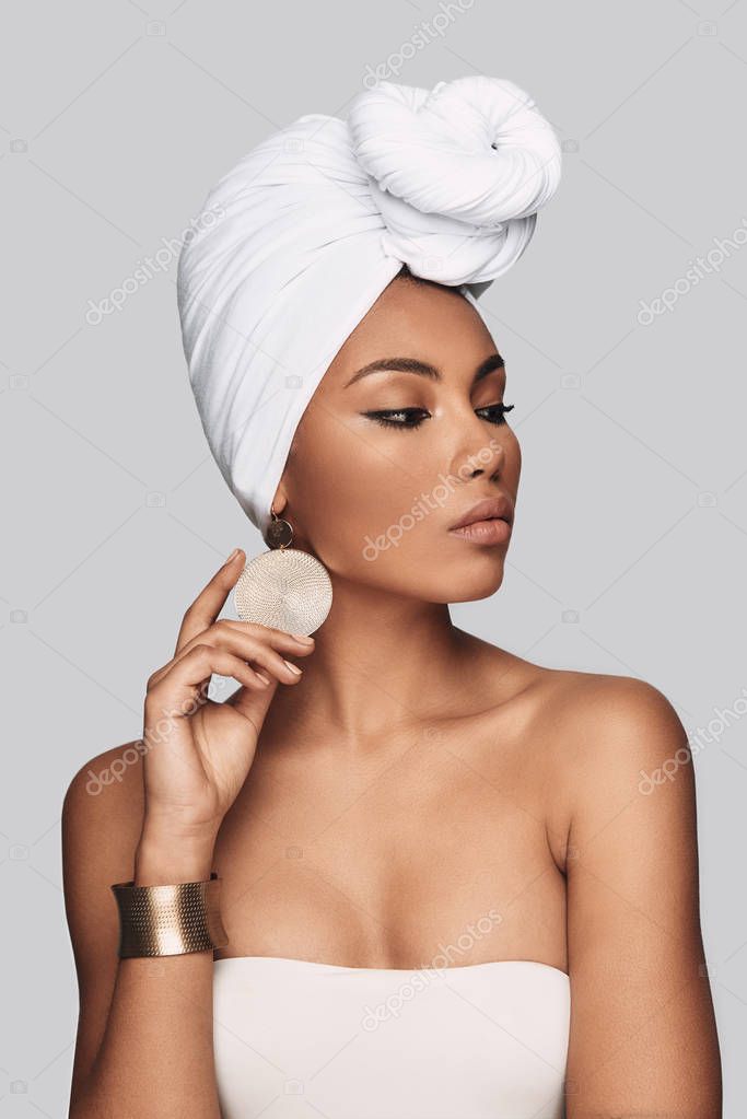 Dangerously beautiful. Attractive young African woman in turban adjusting earring and looking away while standing against grey background
