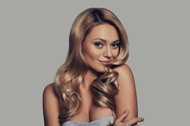 gülümseyen ve kamera gri arka plan karşı dururken bakarak Uzun Kıvırcık sarışın çekici genç kadın