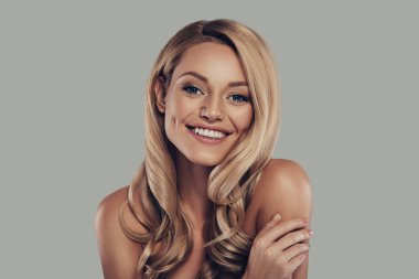 Sakin ve rahat gülümseyen kadın gülümseyerek ve kamera gri arka plan karşı dururken bakarak uzun kıvırcık sarı saçları ile