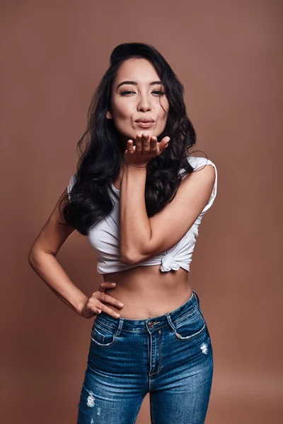 Kiss for you, Beautiful young woman blowing a kiss and smiling while standing against brown background