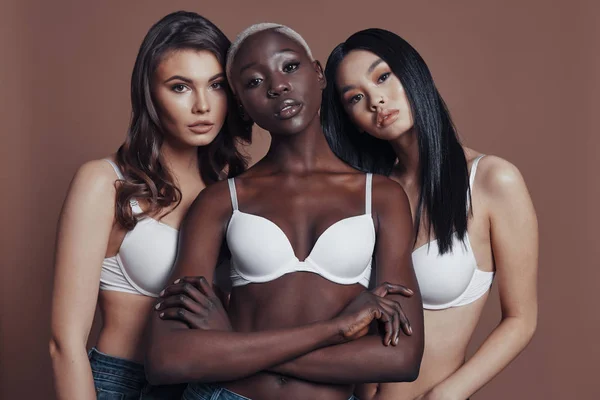 three women wearing bras and posing against brown background and looking at camera