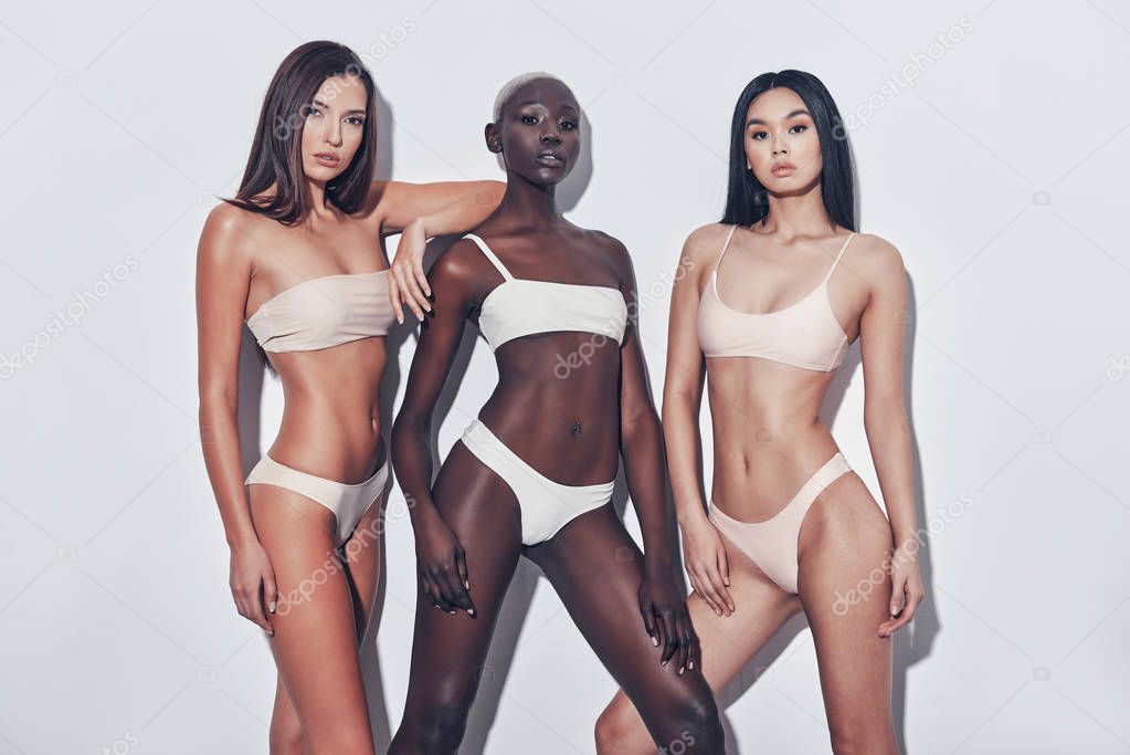 Three attractive mixed race women looking at camera while standing against grey background and wearing lingerie